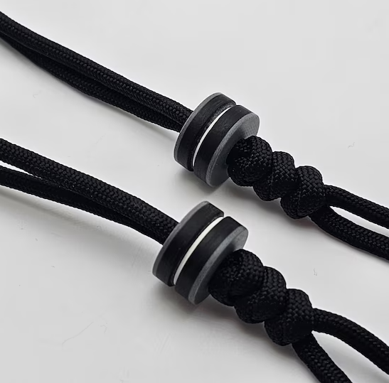 Close-up comparison of G10 bead versus brass and titanium beads on paracord demonstrating weight difference, layered color patterns, and corrosion-resistant surface finish